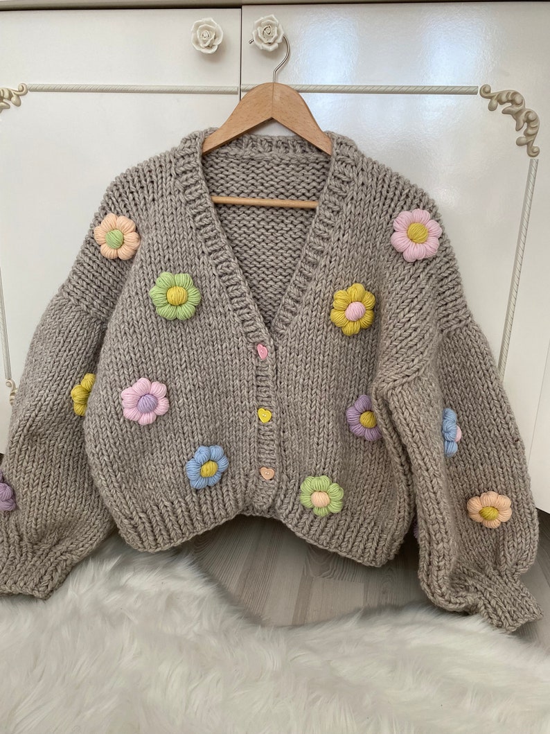 May include: A beige knitted cardigan with a V-neck and long sleeves. The cardigan is decorated with colorful flower appliques.
