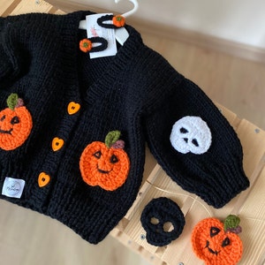 May include: Black knitted cardigan with orange and green pumpkin appliques and white skull applique. The cardigan has heart-shaped buttons and comes with a set of orange pumpkin and white skull hair clips.