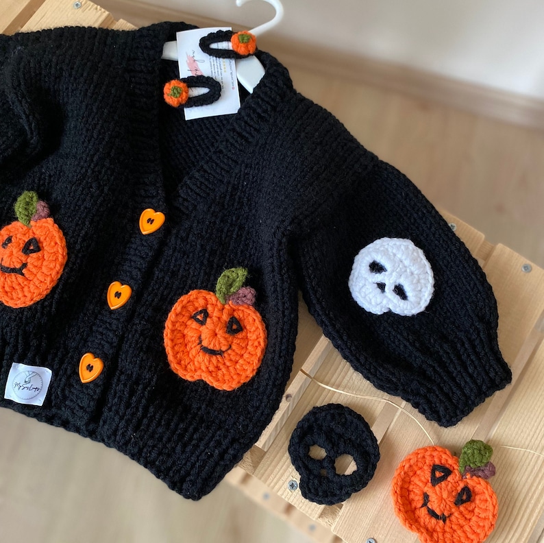 May include: Black knitted cardigan with orange and green pumpkin appliques and white skull applique. The cardigan has heart-shaped buttons and comes with a set of orange pumpkin and white skull hair clips.