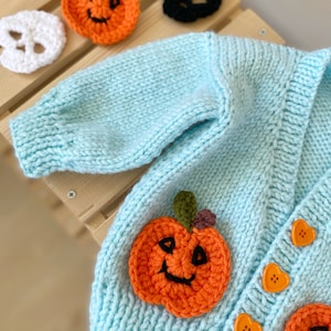 May include: A light blue knitted cardigan with two orange pumpkin appliques and heart-shaped buttons. The cardigan is decorated with two additional pumpkin appliques.