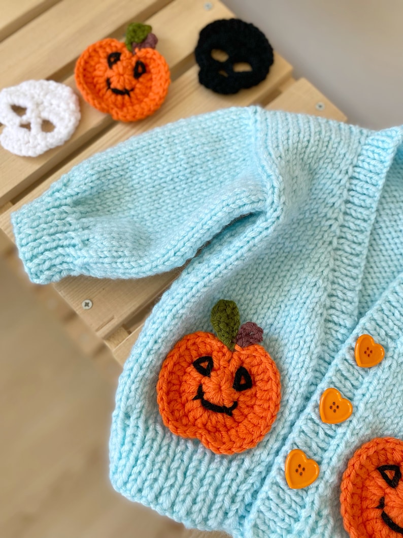 May include: A light blue knitted cardigan with two orange pumpkin appliques and heart-shaped buttons. The cardigan is decorated with two additional pumpkin appliques.