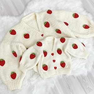 Könnte beinhalten: Zwei weiße Strickjacken mit Erdbeer-Stickerei. Die größere Jacke hat lange Ärmel und einen V-Ausschnitt. Die kleinere Jacke hat kurze Ärmel und einen V-Ausschnitt.