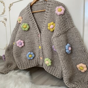 May include: A beige knitted cardigan with a V-neck and button closure. The cardigan is decorated with colorful felt flowers.