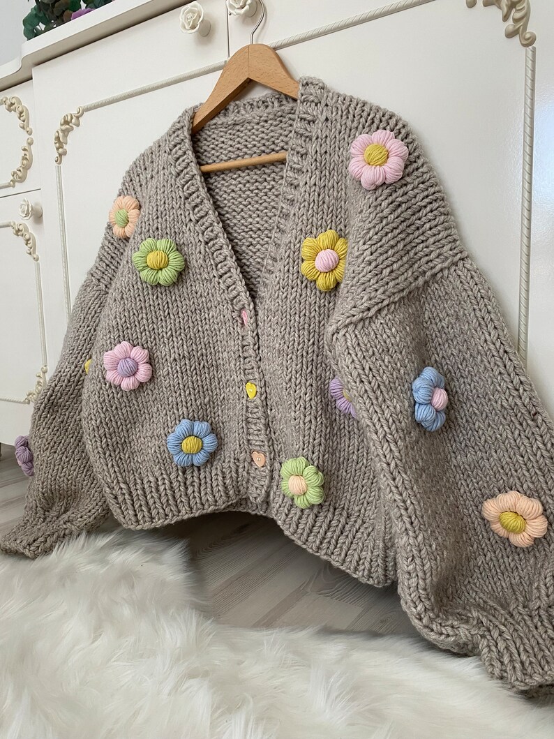 May include: A beige knitted cardigan with a V-neck and button closure. The cardigan is decorated with colorful felt flowers.