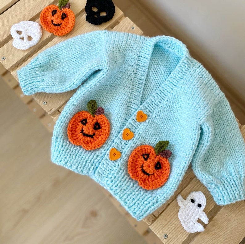 May include: A light blue knitted baby cardigan with pumpkin and ghost appliques. The cardigan has a V-neck and buttons down the front.