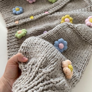 May include: A gray knitted cardigan with a cable knit pattern and colorful flower embellishments.