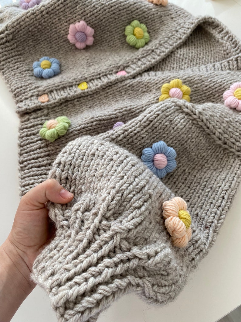 May include: A gray knitted cardigan with a cable knit pattern and colorful flower embellishments.