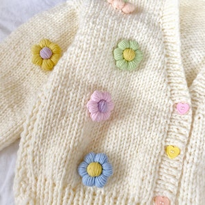 May include: A white knitted cardigan with a cable knit pattern and colorful flower embellishments. The cardigan has a V-neck and heart-shaped buttons.