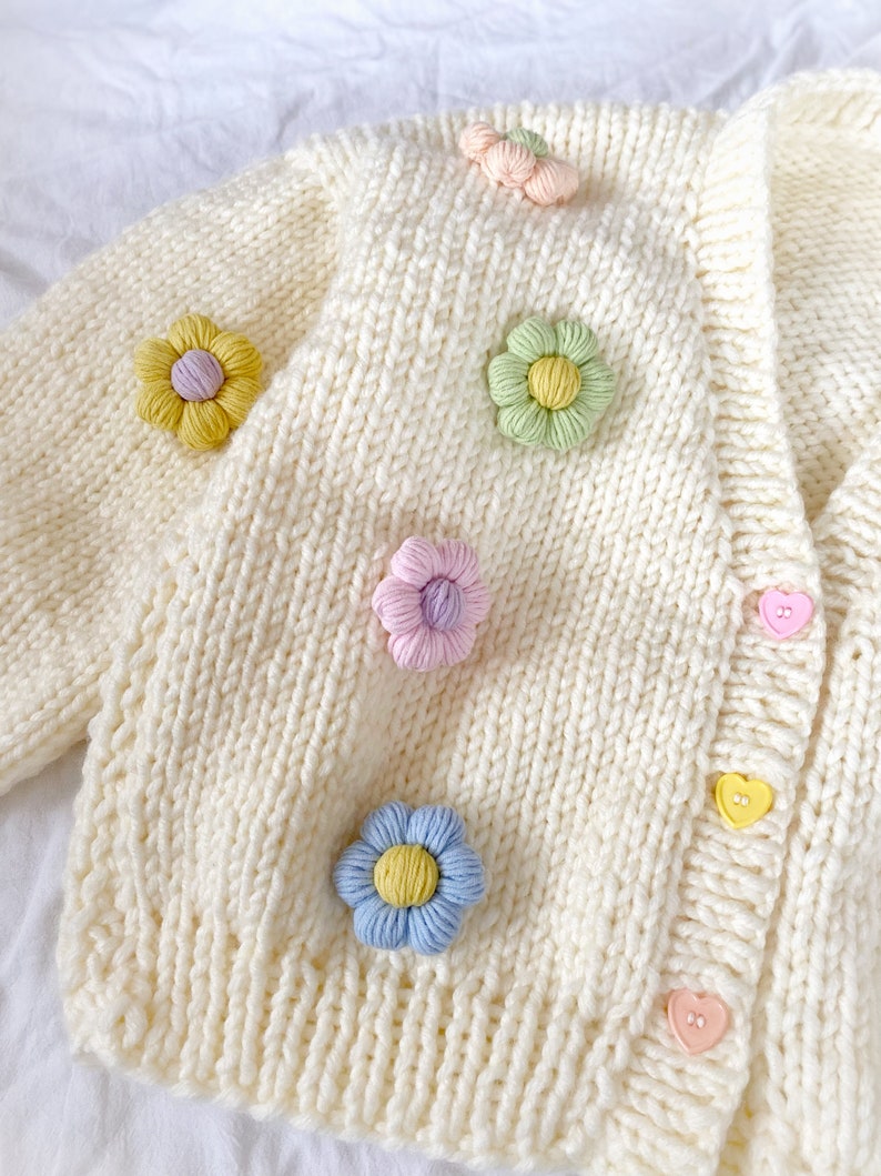 May include: A white knitted cardigan with a cable knit pattern and colorful flower embellishments. The cardigan has a V-neck and heart-shaped buttons.