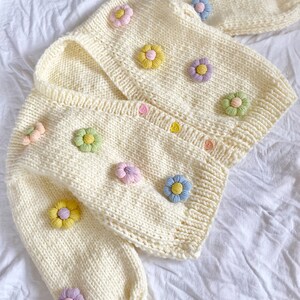 May include: A cream-colored knitted cardigan with a V-neck and long sleeves. The cardigan is decorated with colorful flower appliques.