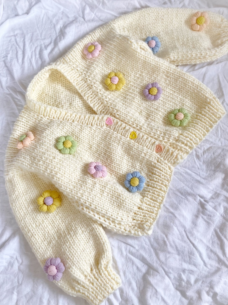 May include: A cream-colored knitted cardigan with a V-neck and long sleeves. The cardigan is decorated with colorful flower appliques.