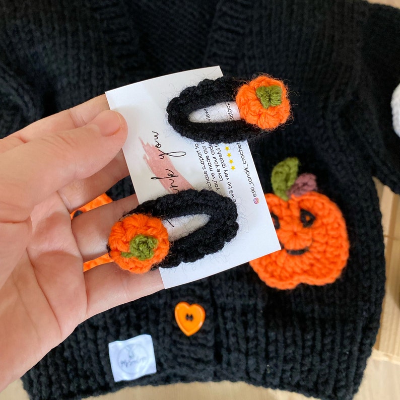May include: Two black and orange crocheted pumpkin hair clips with green stems. The clips are on a white card with the text "Thank you" written in black. The card is held by a hand wearing a black knitted sweater with a crocheted pumpkin patch.
