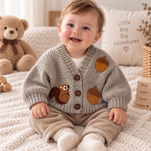 May include: A light grey knitted cardigan featuring three dark brown buttons and embroidered patches of a squirrel and two acorns. The long-sleeved sweater has a ribbed V-neck collar and cuffs, providing a textured look for a young child's outfit.