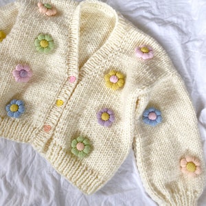 May include: A white knitted cardigan with a V-neck and long sleeves. The cardigan is decorated with colorful crocheted flower embellishments.