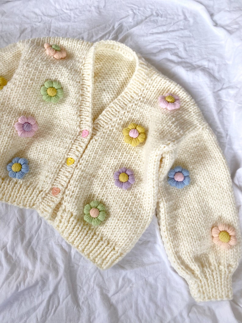 May include: A white knitted cardigan with a V-neck and long sleeves. The cardigan is decorated with colorful crocheted flower embellishments.