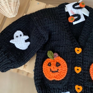 May include: Black knitted cardigan featuring a white ghost, orange pumpkin, and heart-shaped buttons. The cardigan has a "My Salon" tag attached.