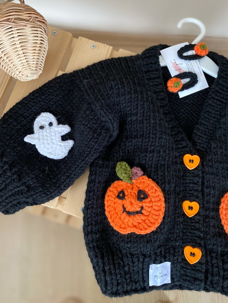 May include: Black knitted cardigan featuring a white ghost, orange pumpkin, and heart-shaped buttons. The cardigan has a "My Salon" tag attached.