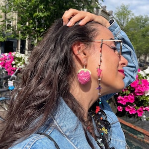 Op de afbeelding: Een vrouw met lang donker haar draagt een spijkerjack en een bril met een kralenketting. De ketting heeft kleurrijke kralen en bedels, waaronder een roze aardbei-bedel die aan een oor hangt.