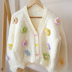 May include: A white knitted cardigan with a V-neck and button closure. The cardigan is decorated with colorful crocheted flower embellishments.