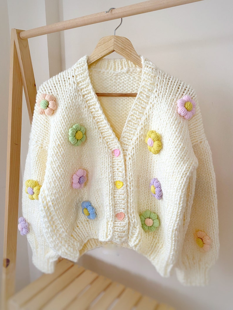 May include: A white knitted cardigan with a V-neck and button closure. The cardigan is decorated with colorful crocheted flower embellishments.