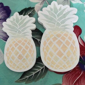 Puede incluir: Dos pegatinas de piña blancas con un diseño verde claro y amarillo. Las piñas están sobre un fondo de tela floral.