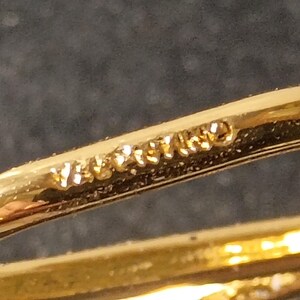 Pu&ograve; includere: Primo piano di un anello dorato con un design curvo. L'anello ha una finitura lucida e la parola "VERITABLE" &egrave; stampata sulla superficie. L'anello &egrave; posto su uno sfondo scuro.