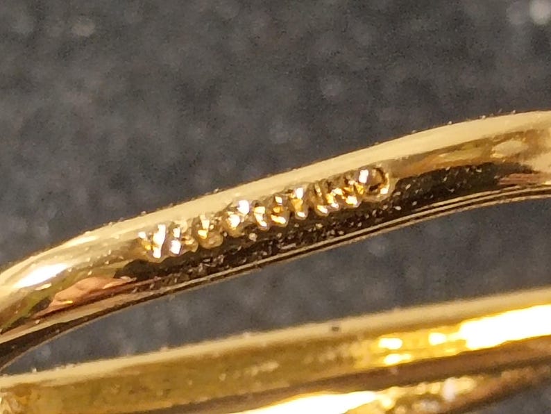 Pu&ograve; includere: Primo piano di un anello dorato con un design curvo. L'anello ha una finitura lucida e la parola "VERITABLE" &egrave; stampata sulla superficie. L'anello &egrave; posto su uno sfondo scuro.