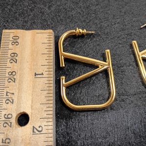 Pu&ograve; includere: Orecchini in metallo dorato a forma delle lettere "V" e "J". Gli orecchini sono lunghi circa 2,5 cm. Gli orecchini sono su una superficie scura accanto a un righello di legno.