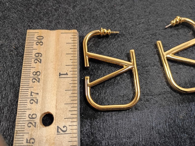 Pu&ograve; includere: Orecchini in metallo dorato a forma delle lettere "V" e "J". Gli orecchini sono lunghi circa 2,5 cm. Gli orecchini sono su una superficie scura accanto a un righello di legno.