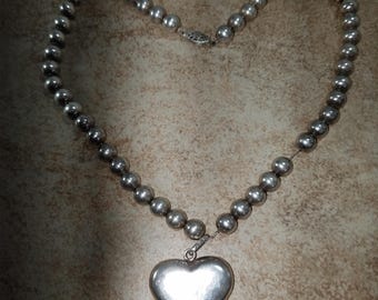 May include: A silver necklace with a large heart pendant. The necklace is made of round beads and has a simple clasp.