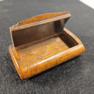 May include: A small, open wooden box with a hinged lid. The box is rectangular with rounded edges and a warm, brown color. The interior is a darker shade of brown. The box appears to be antique or vintage.