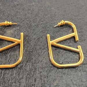 Pu&ograve; includere: Un paio di orecchini a cerchio color oro. Ogni orecchino presenta un design stilizzato a forma di lettera "V". Gli orecchini hanno una chiusura a perno e una finitura lucida, che riflette la luce. Gli orecchini sono esposti su una superficie scura.