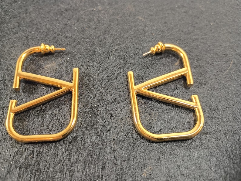 Pu&ograve; includere: Un paio di orecchini a cerchio color oro. Ogni orecchino presenta un design stilizzato a forma di lettera "V". Gli orecchini hanno una chiusura a perno e una finitura lucida, che riflette la luce. Gli orecchini sono esposti su una superficie scura.