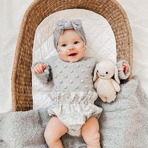 May include: A baby in a grey knitted sweater with bobble detailing, floral bloomers, and a matching bow headband. A stuffed bunny toy is next to the baby in a woven basket. The baby is lying on a grey blanket.