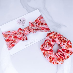 May include: A pink and red floral headband and scrunchie set. The headband is tied in a knot and the scrunchie is made of a soft, stretchy fabric.