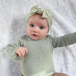 May include: A baby wearing a sage green long-sleeved bodysuit with a floral pattern and a sage green knotted headband.