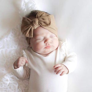 May include: A baby wearing a white long-sleeved onesie with a brown knotted headband. The baby is sleeping on a white lace surface.