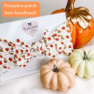 May include: A white fabric headband with a large bow featuring a pumpkin patch print. The bow is tied in a knot and the headband is displayed on a white background with small pumpkins.
