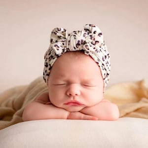 Op de afbeelding: Een pasgeboren baby met een cr&egrave;mekleurige tulband-stijl hoofdband met een zwart- en bruin luipaardmotief. De baby ligt met gesloten ogen en rust met het hoofd op de armen.