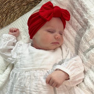 May include: A bright red ribbed fabric headband featuring a large, centred bow detail. The headband is worn on the head of a sleeping infant who is dressed in a long-sleeved white dress with a gathered waist and textured fabric finish.