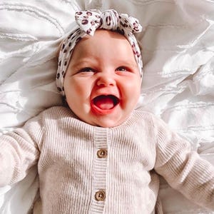 Op de afbeelding: Een lachende baby draagt een beige vest met houten knopen en een haarband met een luipaardprint strik. De baby ligt op een wit, getextureerd oppervlak.