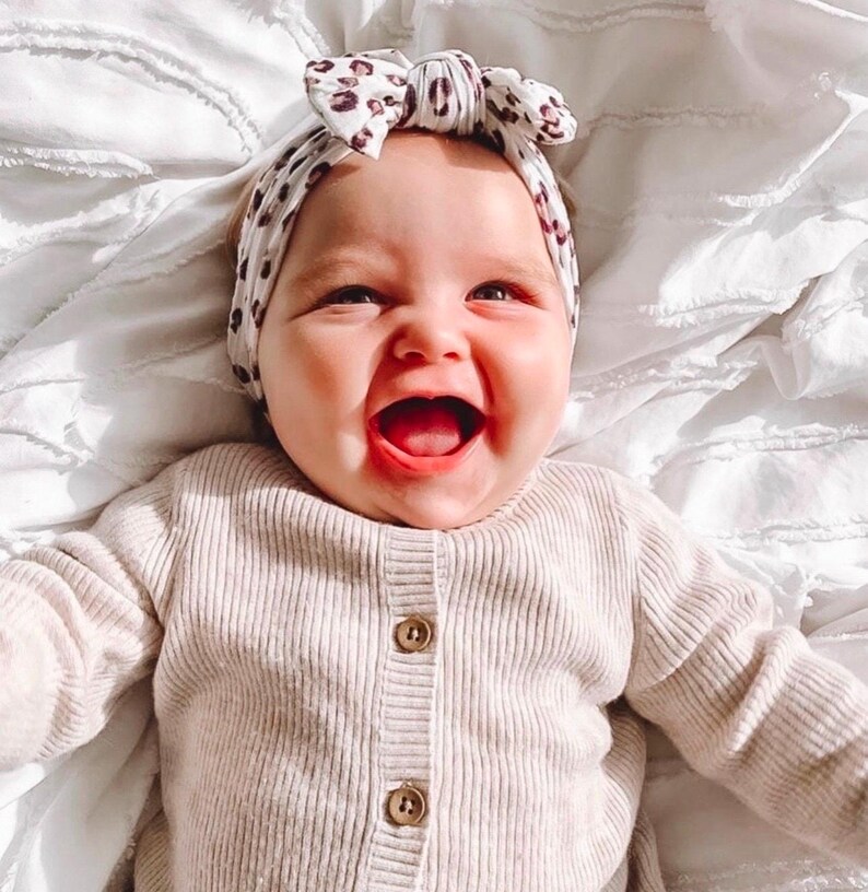 Op de afbeelding: Een lachende baby draagt een beige vest met houten knopen en een haarband met een luipaardprint strik. De baby ligt op een wit, getextureerd oppervlak.