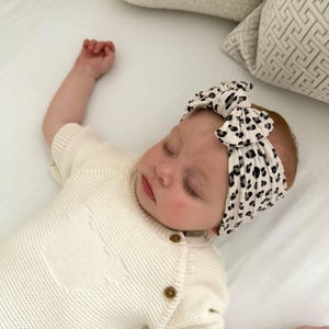 Op de afbeelding: Een slapende baby draagt een cr&egrave;mekleurige gebreide romper met houten knopen en een zwart-witte luipaardprint hoofdband. De baby ligt op een wit laken met kussens in neutrale tinten op de achtergrond.