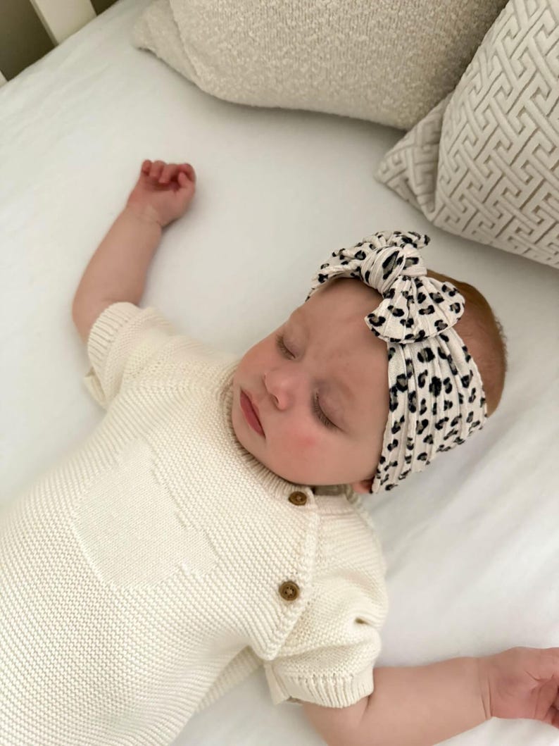 Op de afbeelding: Een slapende baby draagt een cr&egrave;mekleurige gebreide romper met houten knopen en een zwart-witte luipaardprint hoofdband. De baby ligt op een wit laken met kussens in neutrale tinten op de achtergrond.