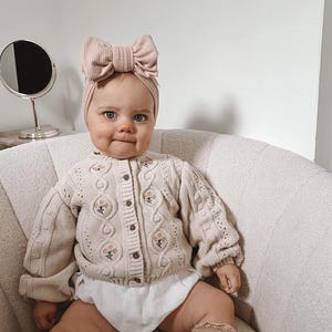 May include: A baby in a light beige cable knit cardigan with floral embroidery and wooden buttons. The baby also wears a matching bow headband and white diaper cover. The baby is seated in a cream-coloured chair.