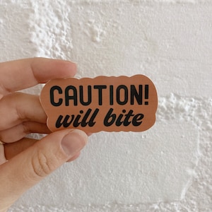 Puede incluir: Una pegatina marrón con el texto "CAUTION! will bite" en letras negras.