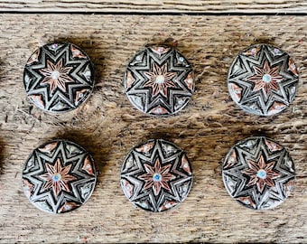 May include: Ten silver and copper star-shaped buttons with a clear crystal center. The buttons have a geometric design and are arranged in two rows on a wooden surface.