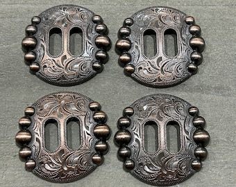 May include: Four antique silver-toned metal conchos with a decorative floral pattern and a double-slot design. Each concho has a circle of small beads around the edge.