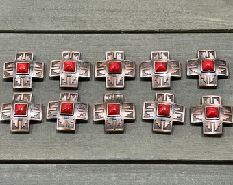 May include: Ten copper-coloured metal conchos with red stone inlays. Each concho is shaped like a cross with intricate designs.