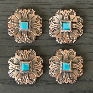May include: Four silver-toned metal conchos with turquoise blue square stones in the center. Each concho has a decorative, ornate design.
